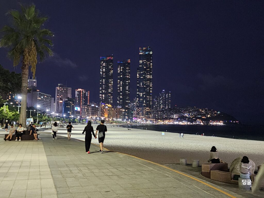 People strolling along the lit boardwalk of Haeundae Beach at night with distant city lights.