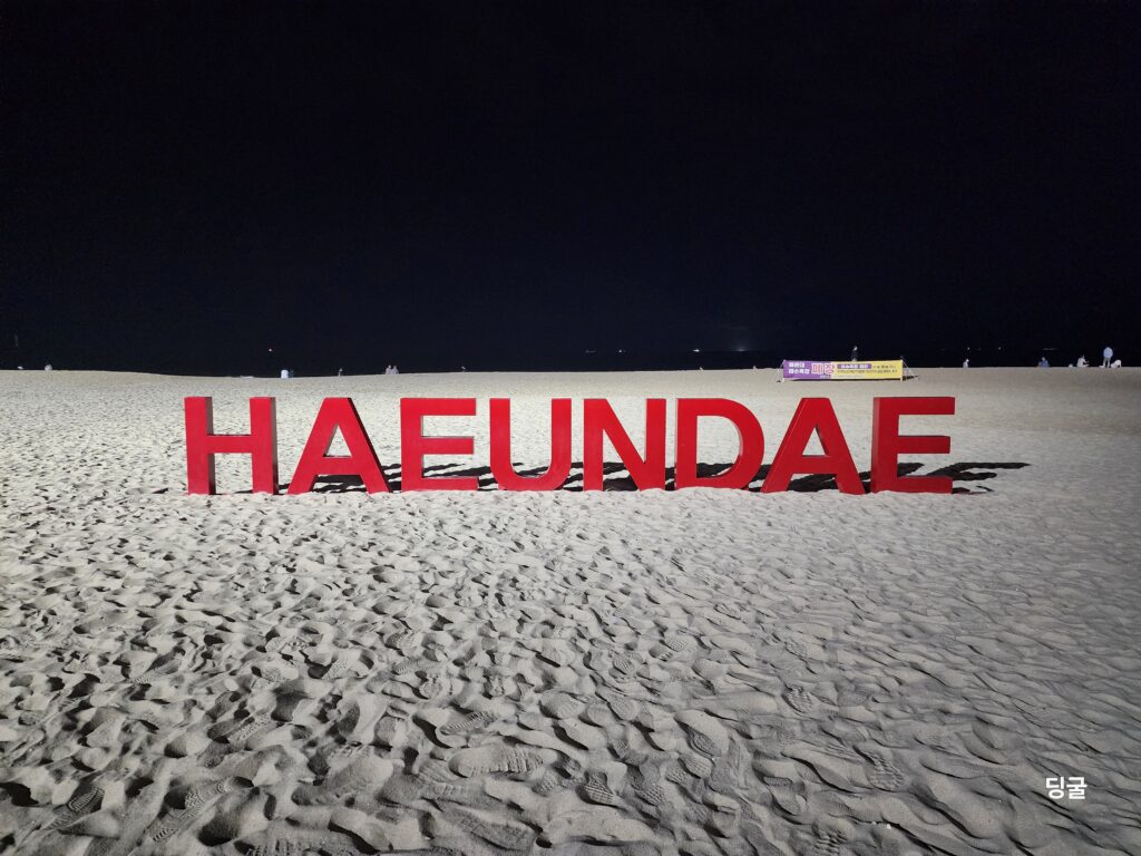 The iconic red HAEUNDAE text sign standing on the white sand under a dark night sky.