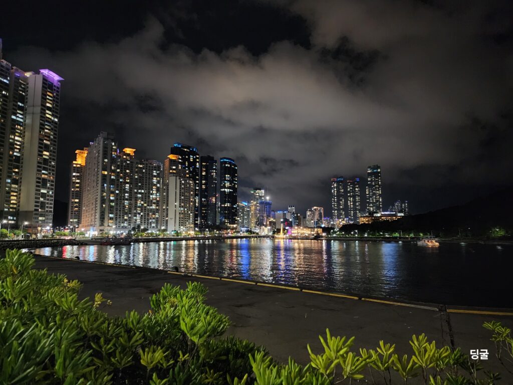 Futuristic night view of high-rise skyscrapers reflecting vibrant neon lights onto the calm water of Haeundae waterfront.
