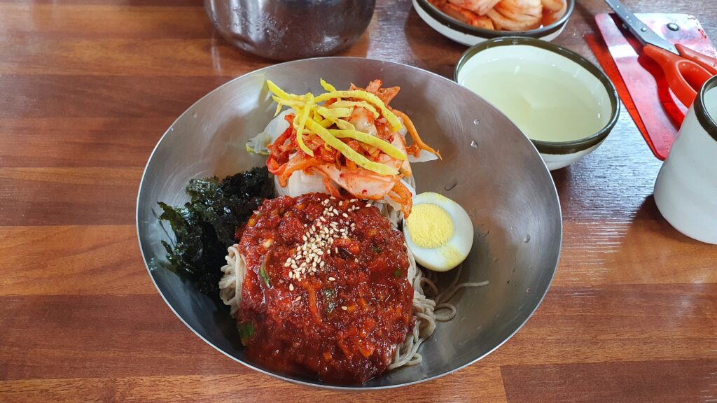 A metal bowl of Korean spicy cold noodles topped with red sauce, sesame seeds, half a boiled egg, dried seaweed, shredded cucumber, and egg garnish.