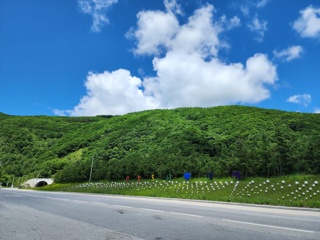 Scenic highway running alongside green mountain slopes under a vibrant blue sky with fluffy clouds.