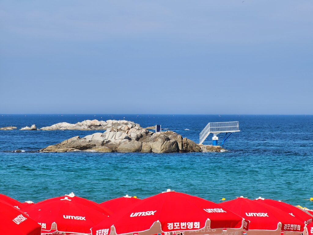 Emerald sea and red beach umbrellas with a rocky island visible in the distance under a blue sky.