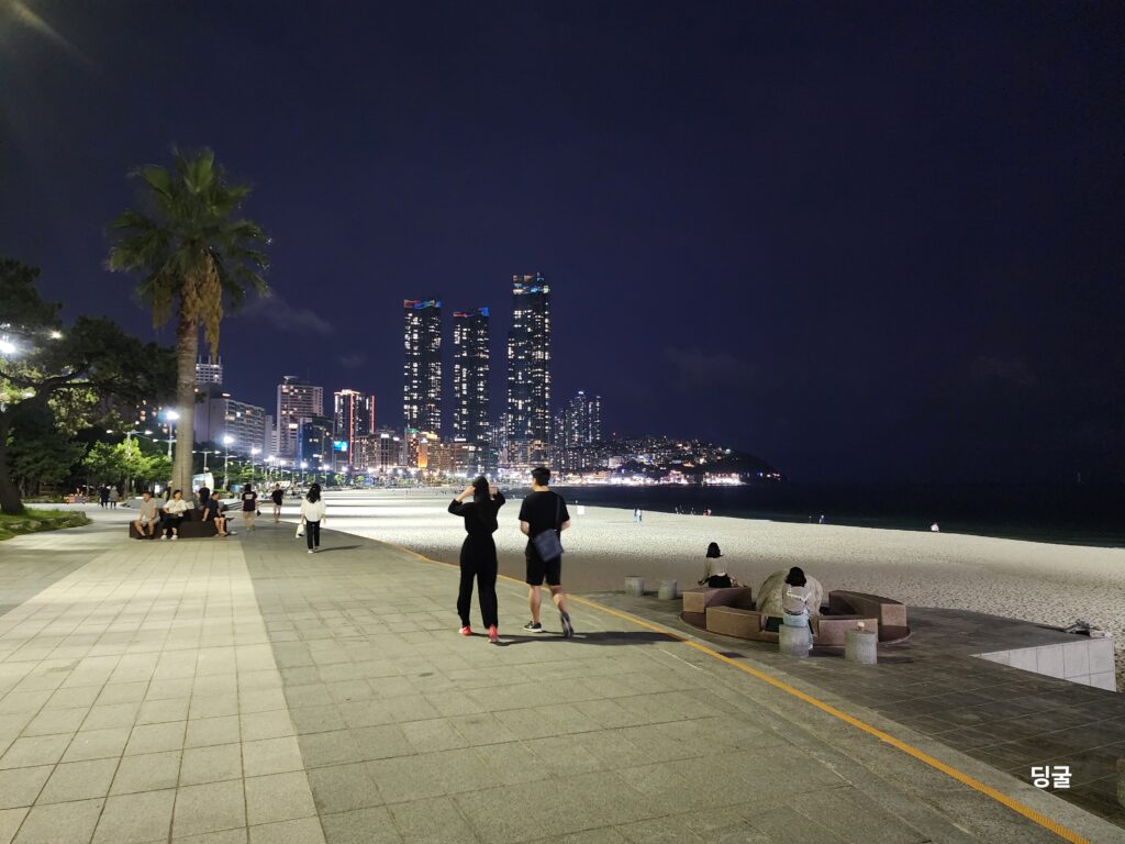 People walking along a paved coastal promenade in Busan at night with distant city lights.
