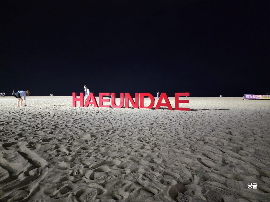 Red 'HAEUNDAE' letter sign standing illuminated on the sandy beach at night.