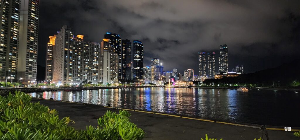 Night view of Busan's coastal skyline with illuminated skyscrapers reflecting on the water under a cloudy sky. cool places to visit in south korea