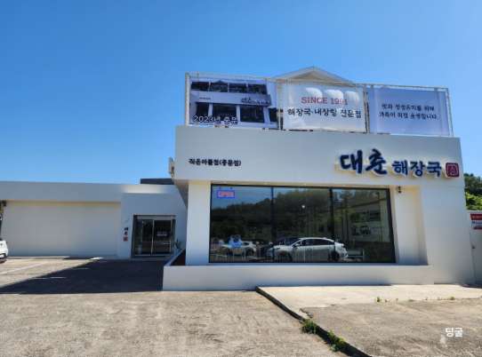 The clean, modern exterior of the Daechun Haejangguk restaurant in Jungmun, Jeju, with a large parking lot.